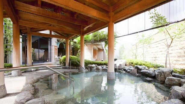 Un onsen japonais traditionnel, avec des marches en pierre menant à un bassin d’eau claire, des poutres en bois au-dessus et un mur de pierre en arrière-plan.