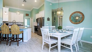 Una cocina moderna con armarios blancos, una isla central con taburetes de bar y un comedor con mesa y sillas blancas.