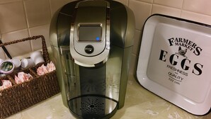 Una cafetera con pantalla digital, un cesto con cápsulas de café y una bandeja con información sobre Huevos de Mercado de Productores.