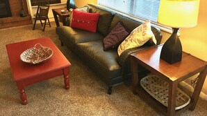 Una sala de estar acogedora con un sofá de cuero, cojines rojos y con diseños, una mesa de centro de madera con un cuenco tejido, una lámpara en una mesita auxiliar y una estantería con diversos objetos.