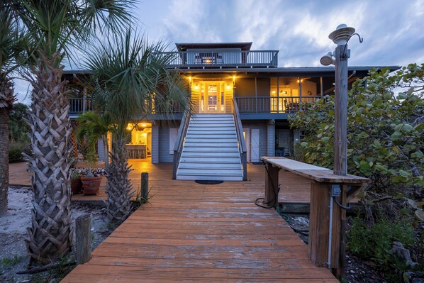 A two-story house with a wooden deck leading to a front door, surrounded by palm trees and a garden.