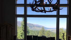 Ein Wohnzimmer mit einem großen Fenster mit Blick auf eine gebirgige Landschaft, einer Deckenleuchte mit Geweihmotiven und einer gemütlichen Sitzecke.