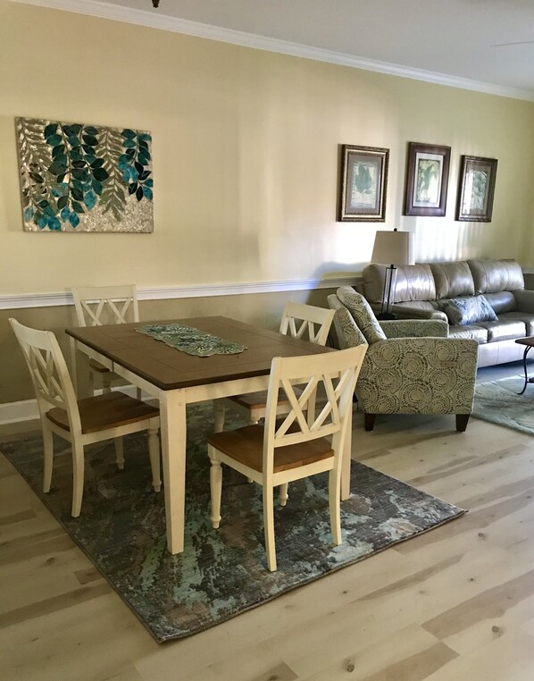 A dining area with a wooden table and chairs, a patterned rug, and a sofa in the background.