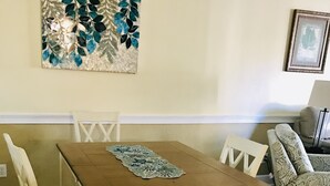 A dining area with a wooden table and white chairs, a patterned rug, and a decorative wall piece. - Beautiful  2B/2B, ground floor condo in Riverwalk on Arrowhead Golf course ! (Myrtle Beach)
