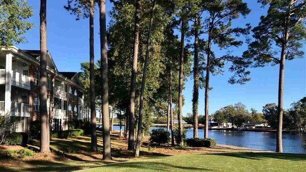 A well-maintained lawn with tall trees and a building with balconies overlooking a body of water. - Beautiful  2B/2B, ground floor condo in Riverwalk on Arrowhead Golf course ! (Myrtle Beach)