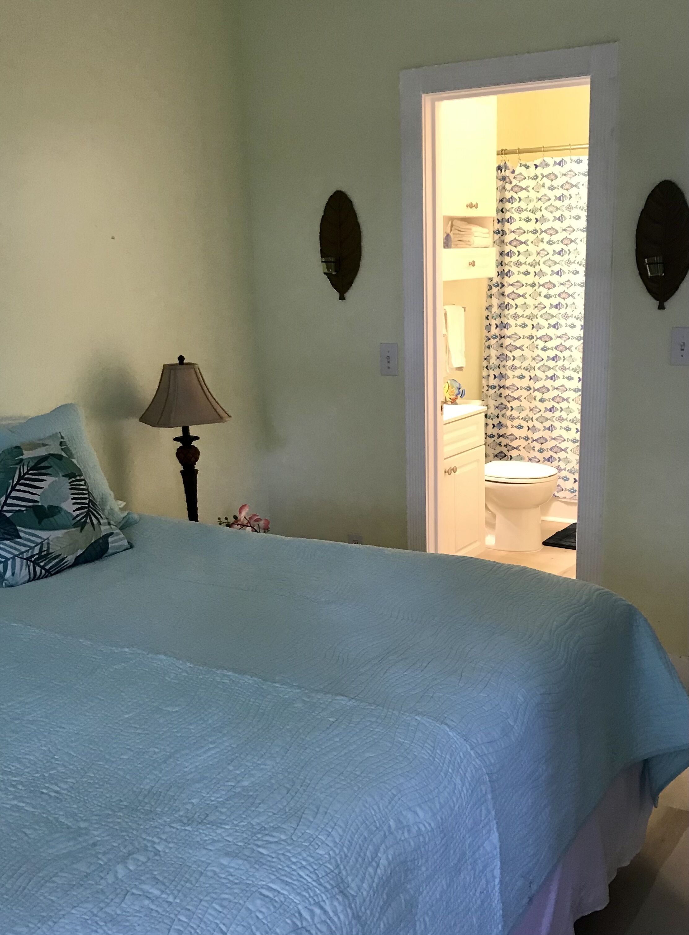 Een slaapkamer met een bed, nachtkastje, lamp en een badkamer door een geopende deur zichtbaar.