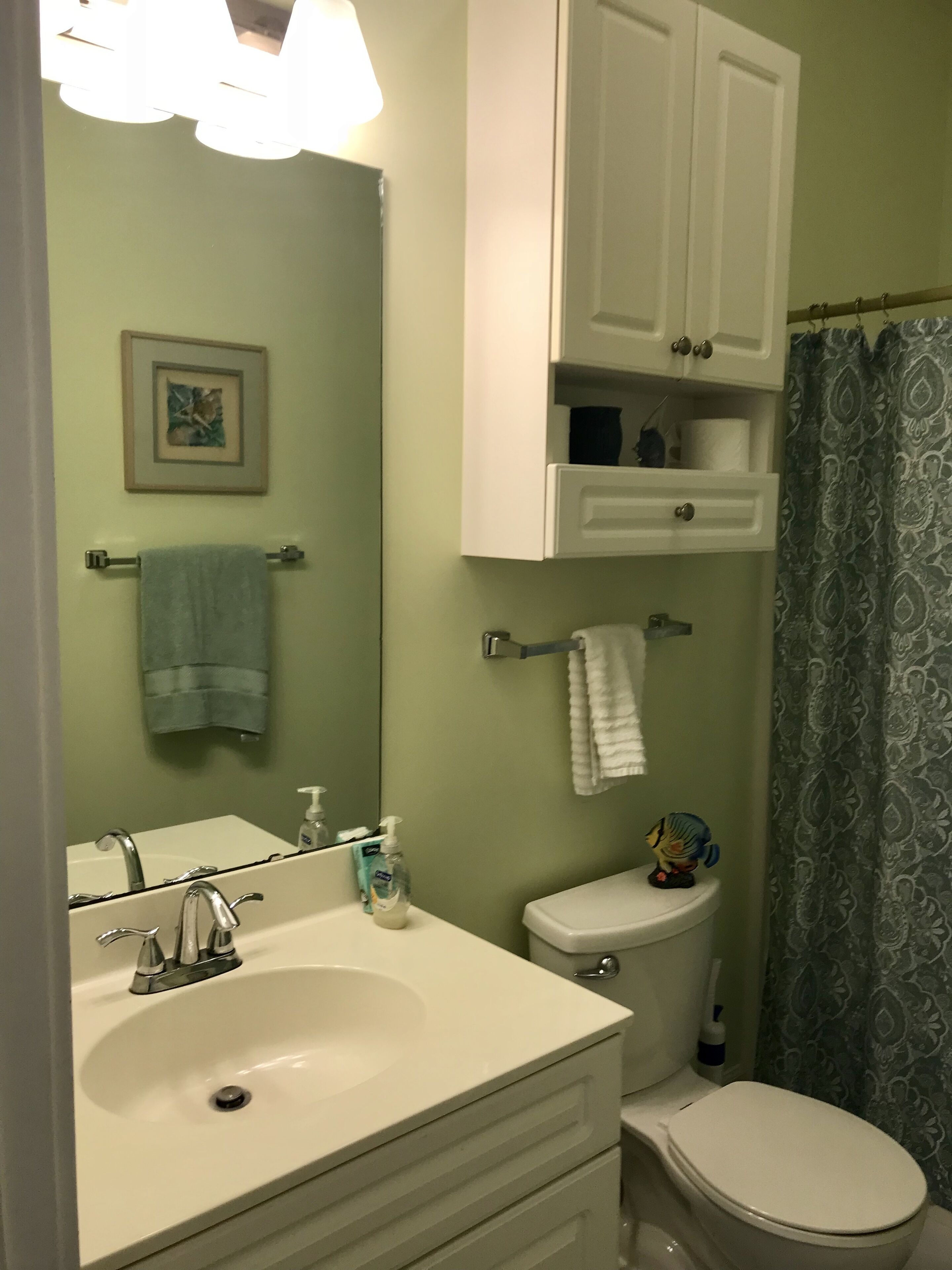 A bathroom with a white sink, mirror, and toilet. There is a towel rack with a towel, a framed picture on the wall, and a shower curtain.