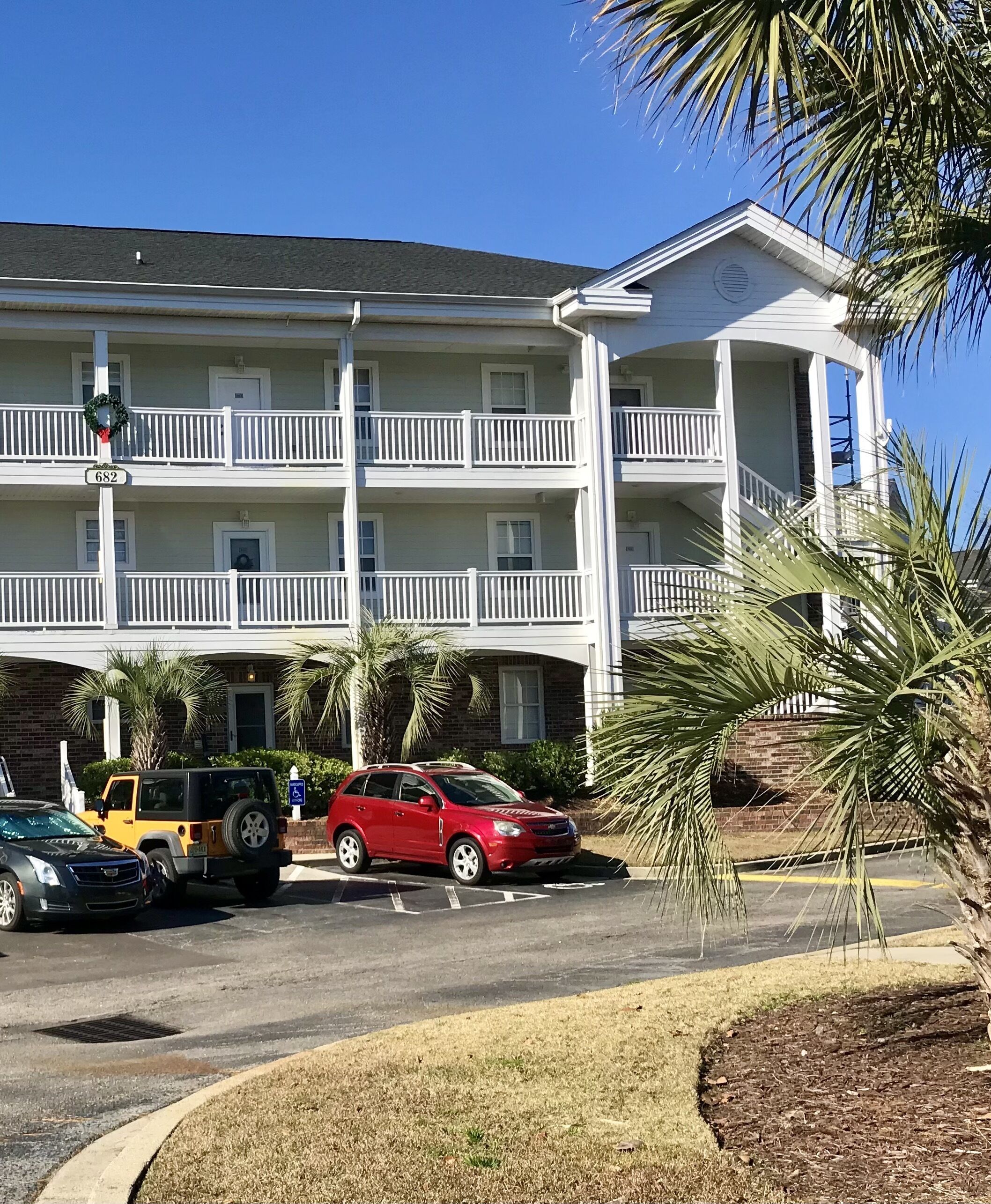 Un hotel a più piani con facciata bianca, un balcone al secondo piano e un parcheggio con un'auto rossa e un veicolo giallo.