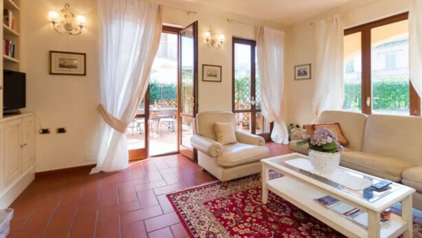 A living room with a sofa, armchair, coffee table, and a TV on a cabinet. There are curtains, a red rug, and a balcony with a table and chairs.