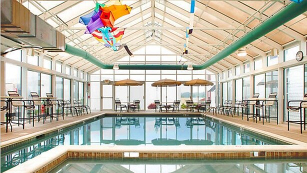 Una amplia piscina interior con paredes de vidrio transparente, rodeada de sillas y mesas de descanso.