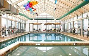 Una amplia piscina interior con paredes de vidrio transparente, rodeada de sillas y mesas de descanso.