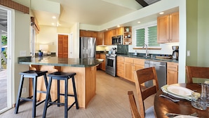 A modern kitchen with wooden cabinets, stainless steel appliances, and a bar stool area. - Hale Ohe | Historic Beachfront Home with A/C + Baby Beach (Koloa)