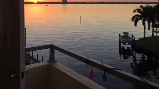 Sonnenuntergangblick von einem Balkon mit ruhigem Gewässer, einem entfernten Gebäude und einer Anlegestelle mit einem Boot.