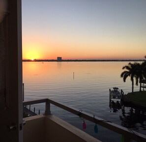 Sonnenuntergangblick von einem Balkon mit ruhigem Gewässer, einem entfernten Gebäude und einer Anlegestelle mit einem Boot.