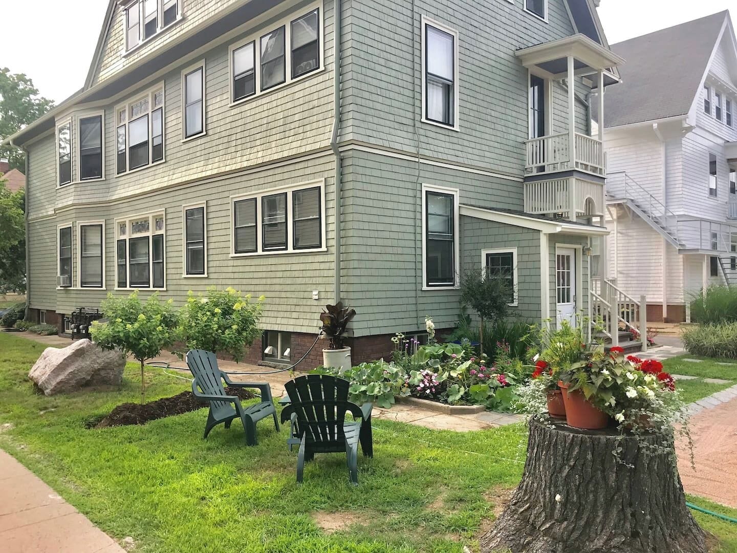 A two-story house with a front porch, a small garden with potted plants, and two Adirondack chairs.