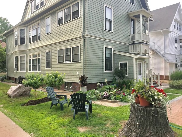 A two-story house with a front porch, a small garden with potted plants, and two Adirondack chairs.