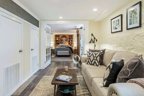 A living room with a sofa, a coffee table, and a mirror. There is a hallway leading to a bedroom with a bed and a wardrobe.