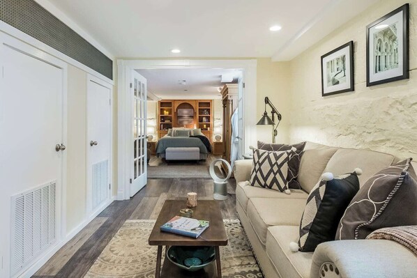 A living room with a sofa, a coffee table, and a mirror. There is a hallway leading to a bedroom with a bed and a wardrobe.