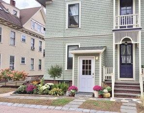 A two-story house with a front porch, a white door, and a blue door. There are potted plants and flowers in front of the house.