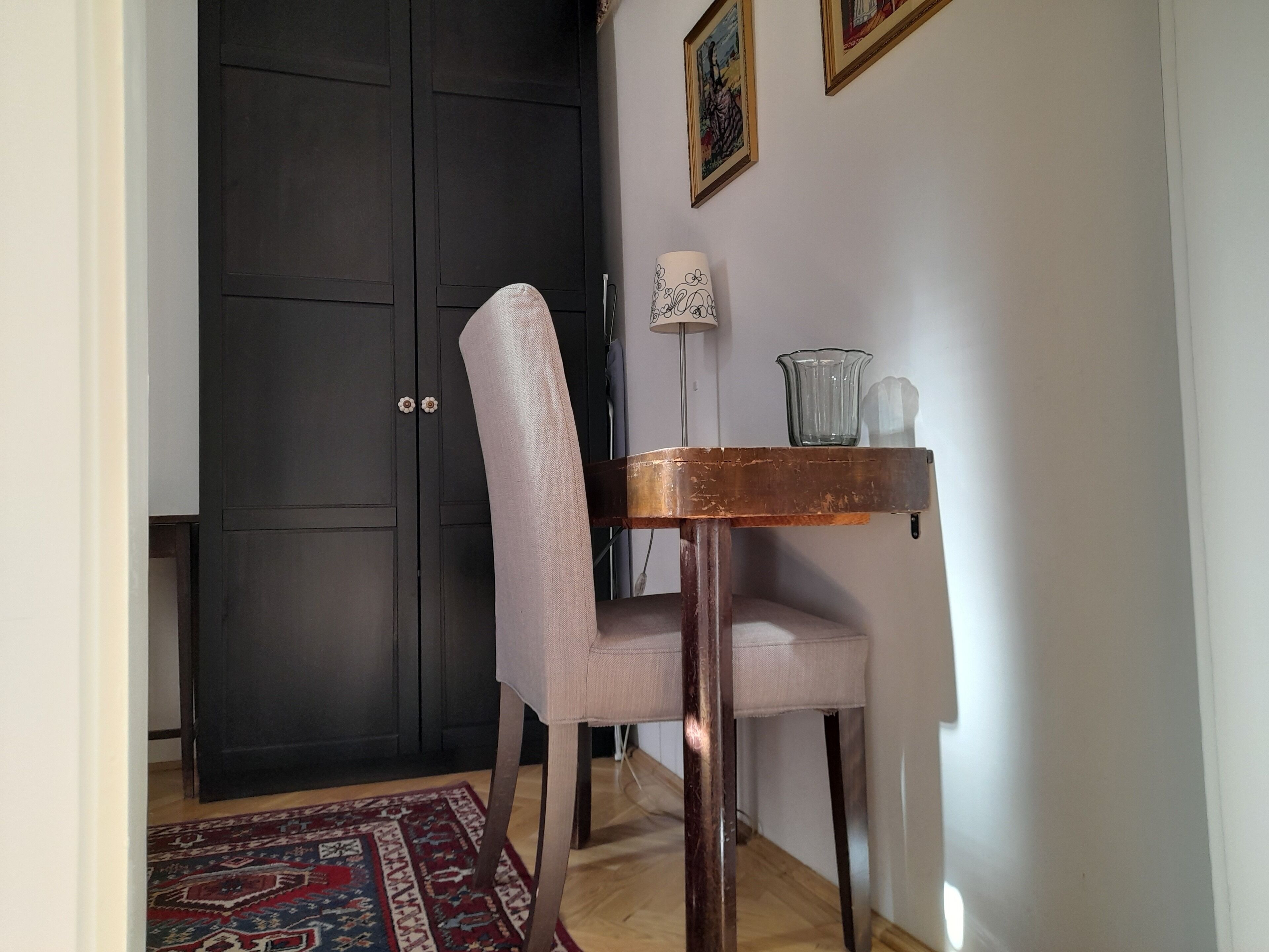 A room with a dark wooden wardrobe, a light-colored chair, a small table with a glass pitcher, and a patterned rug on the floor.