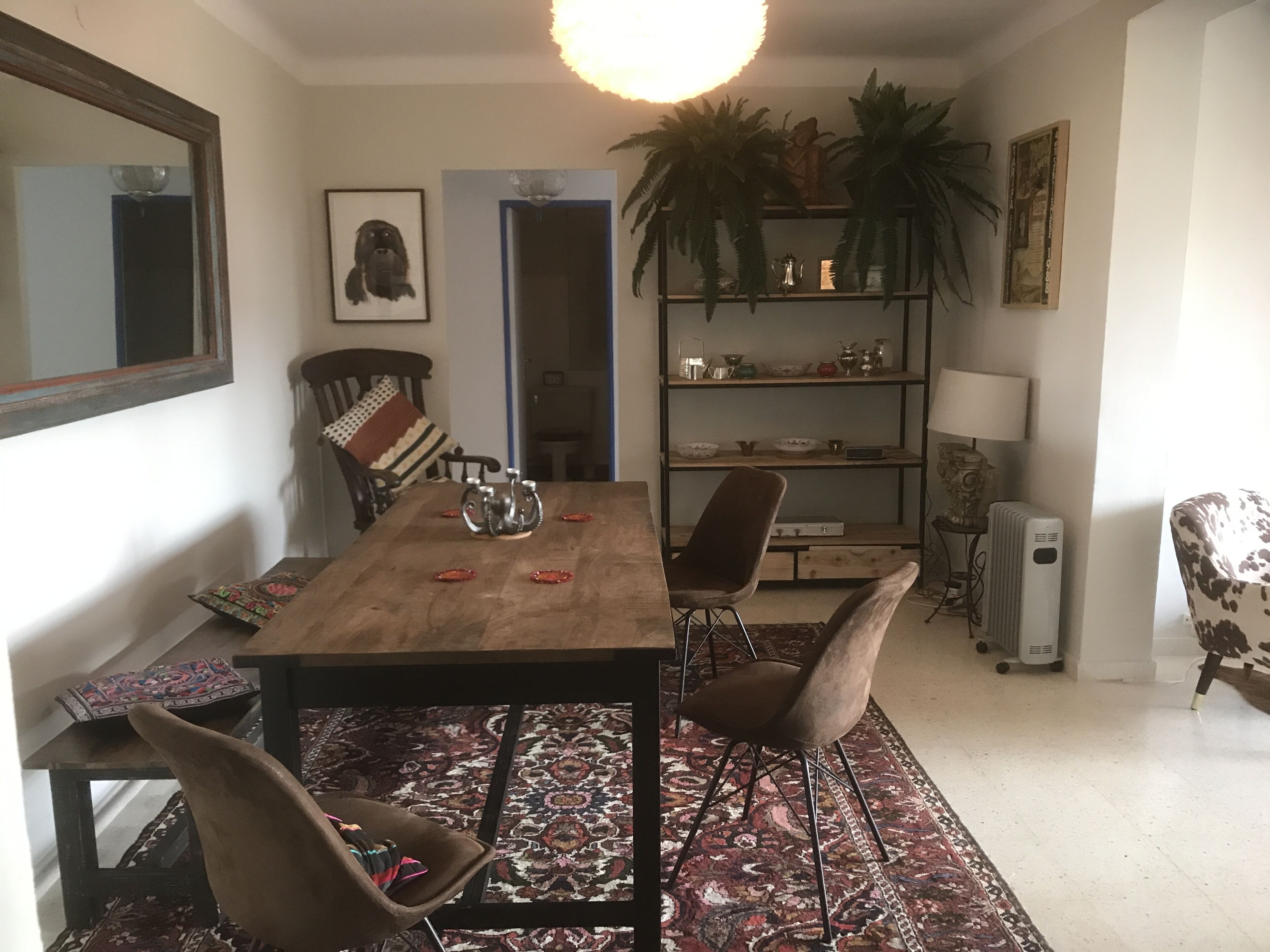A dining area with a wooden table, two chairs, a patterned rug, a bookshelf, and a radiator.