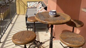 Ein Balkon mit Holztisch und Hockern, eine Sitzecke mit gemusterten Kissen, einer Uhr und Blick auf den Strand.