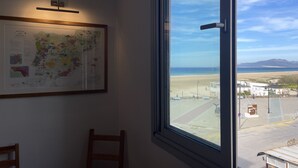 Ein Zimmer mit großem Fenster und Blick auf einen Strand, einer an der Wand hängenden Karte und einem Holzstuhl.