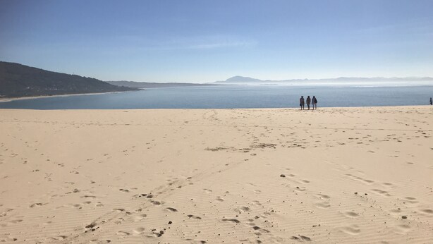 Ein weiter Sandstrand mit Fußspuren, die zum Wasser führen, ein klarer Himmel und ferne Berge.