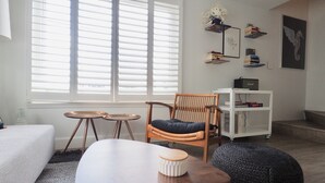 A living room with a large window, a wooden chair, a white sofa, a small round table, and a bookshelf with books and a radio. - Relax near St. Pete & Clearwater Beaches, stay in our 2-bedroom beach condo (Indian Rocks Beach)