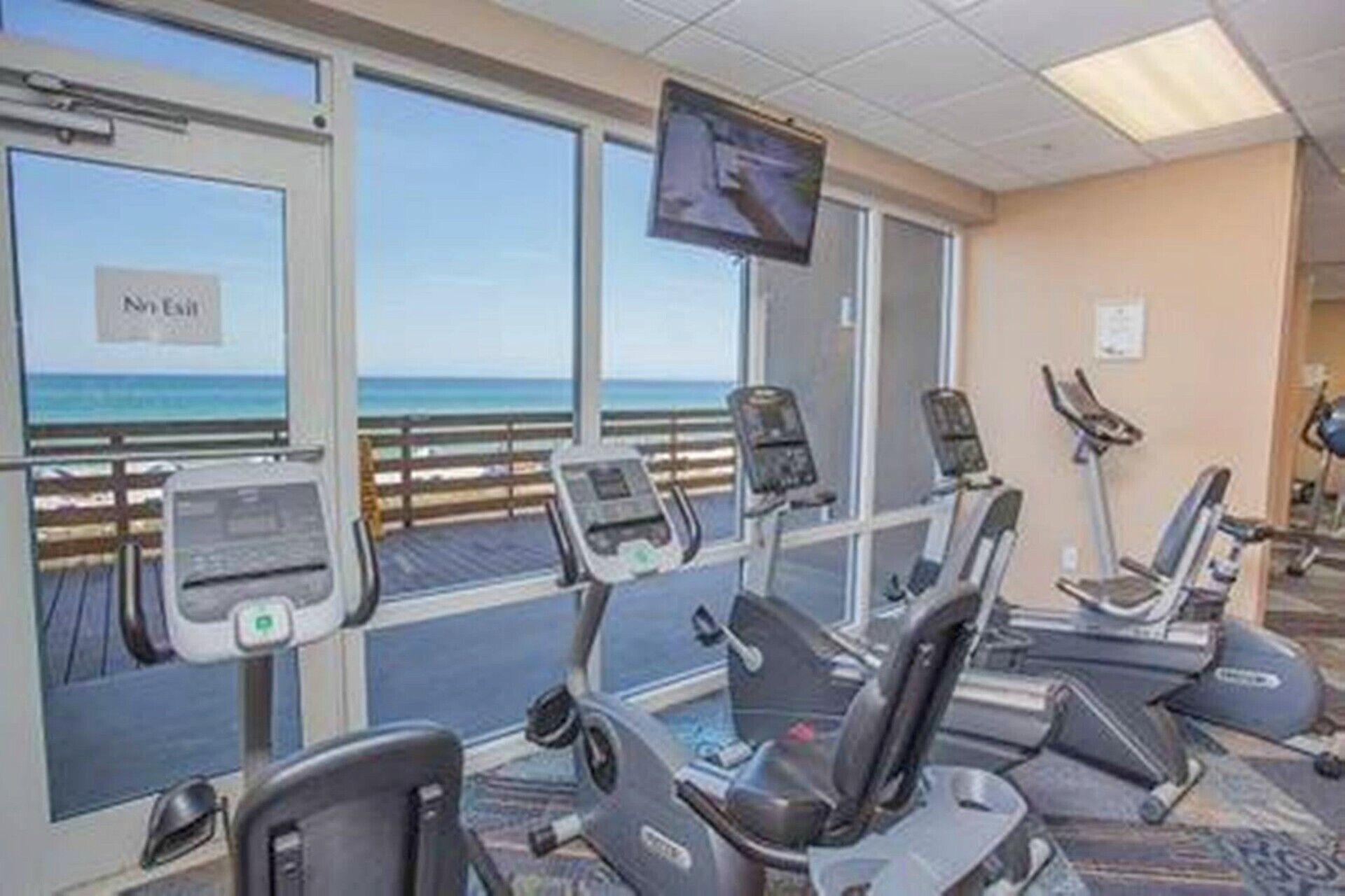 A fitness room with treadmills and exercise equipment, large windows with a view of the beach, and a television mounted on the wall.