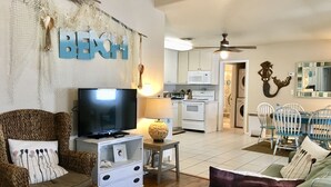 Una sala de estar con televisor, mobiliario de mimbre, una zona de cocina y un ventilador de techo.