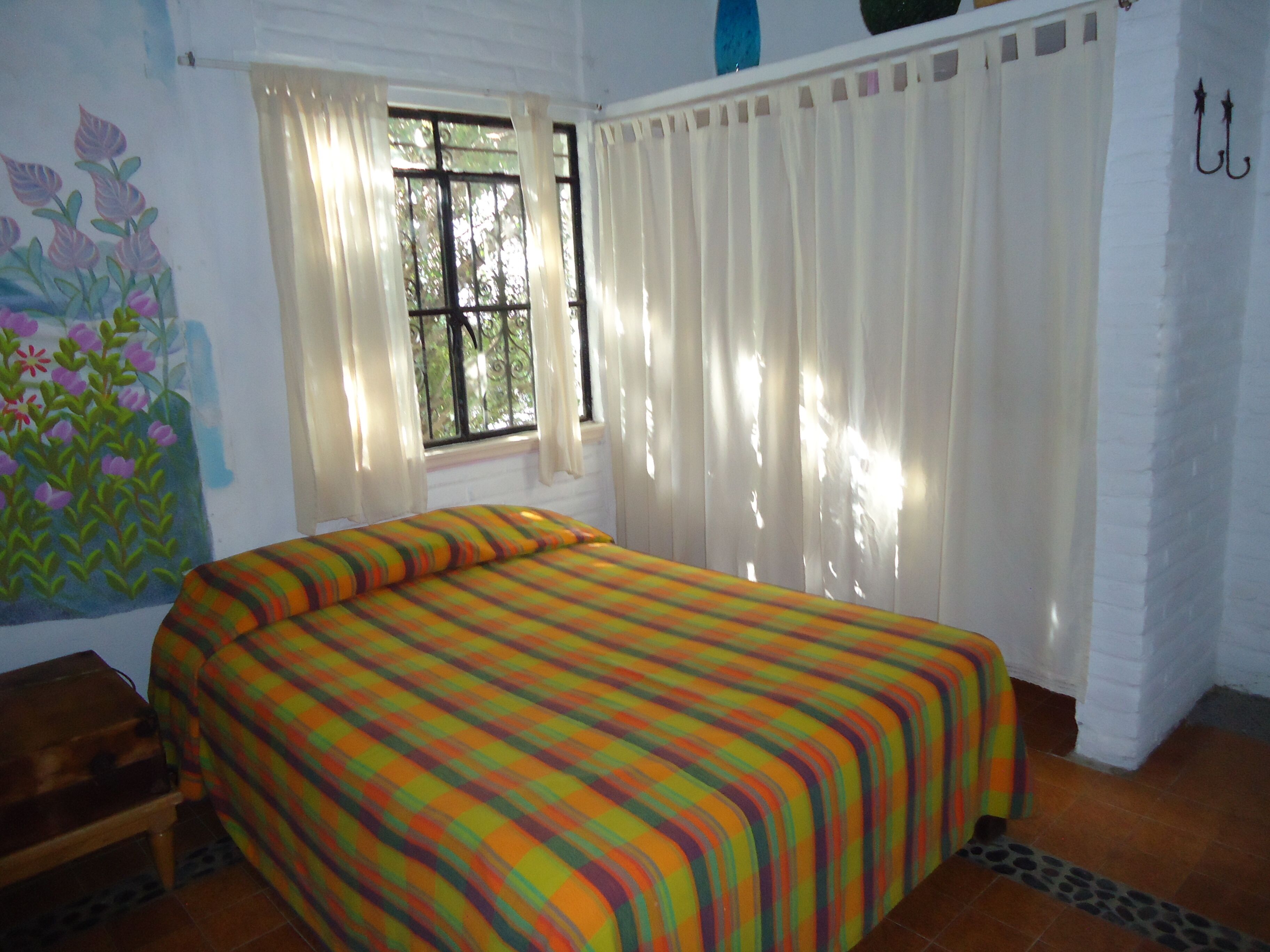 Une pièce comprenant un lit, une fenêtre avec des rideaux, un mur orné d’une fresque florale et une petite table en bois.