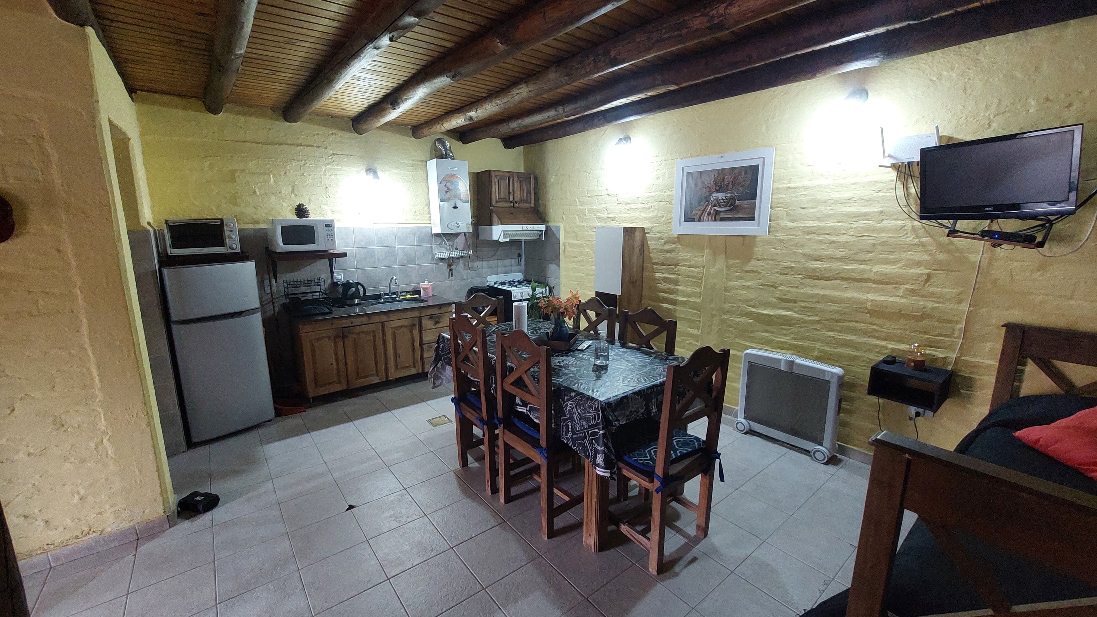 A compact kitchen with wooden cabinets, a dining table with chairs, a microwave, a stove, and a television mounted on the wall.
