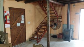 Un escalier en bois avec un ventilateur et un radiateur, une porte en bois ornée de panneaux indiquant les consignes de sécurité incendie, et un sol carrelé.