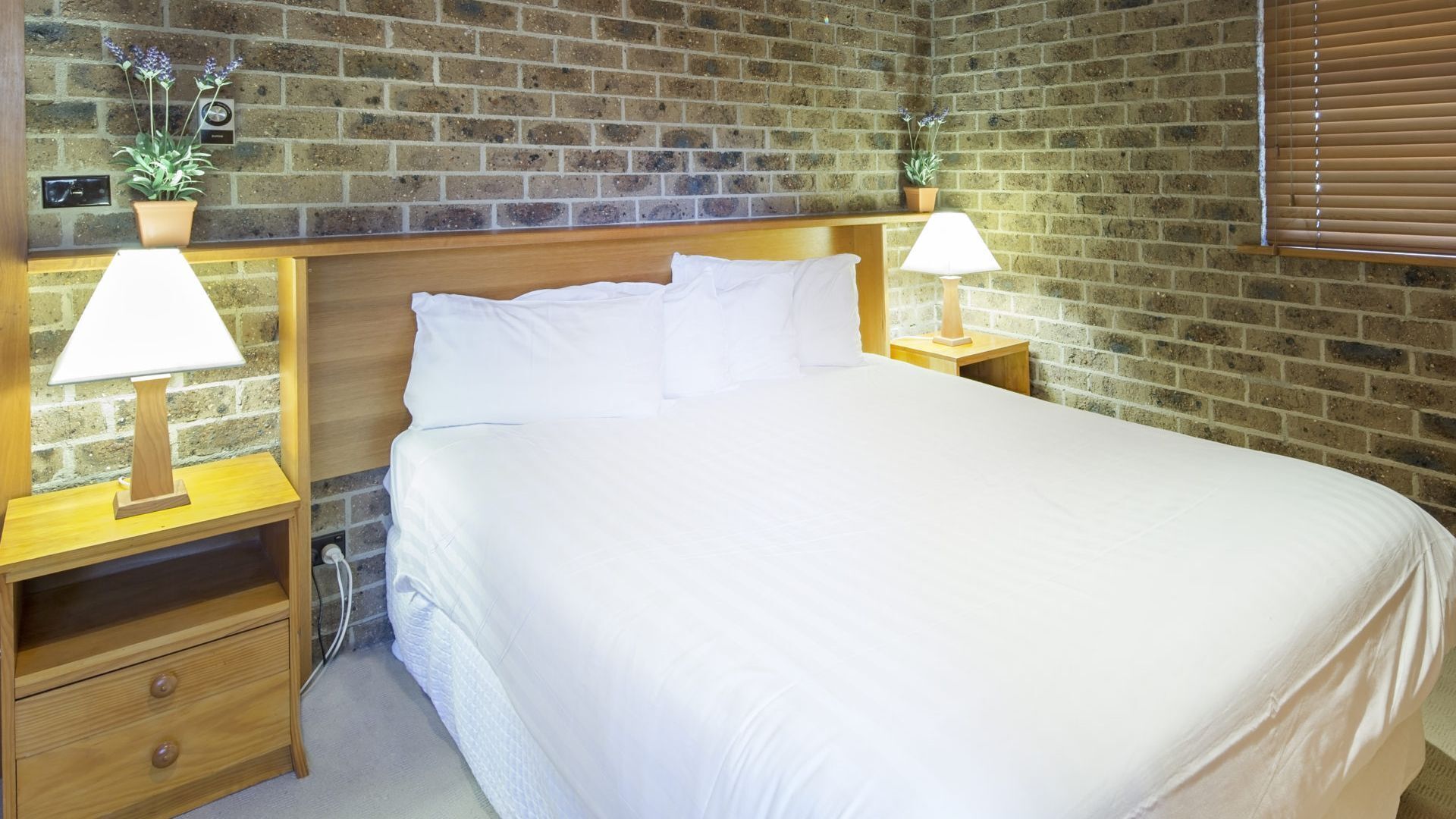A neatly made bed in a room with a brick wall, two bedside tables with lamps, and a small drawer.