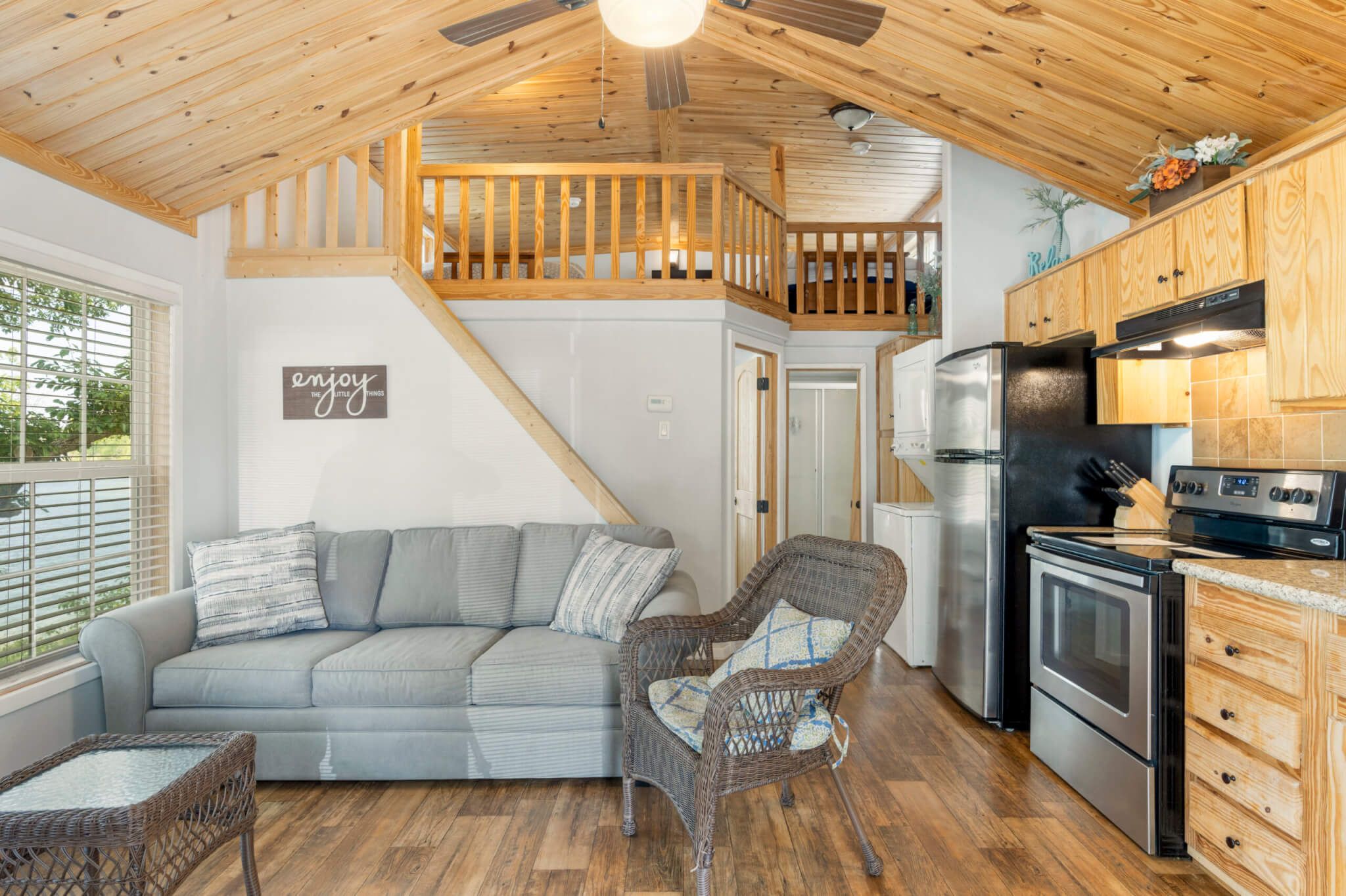 A cozy kitchen with a wooden ceiling, a grey sofa, wicker chairs, and a stainless steel refrigerator.