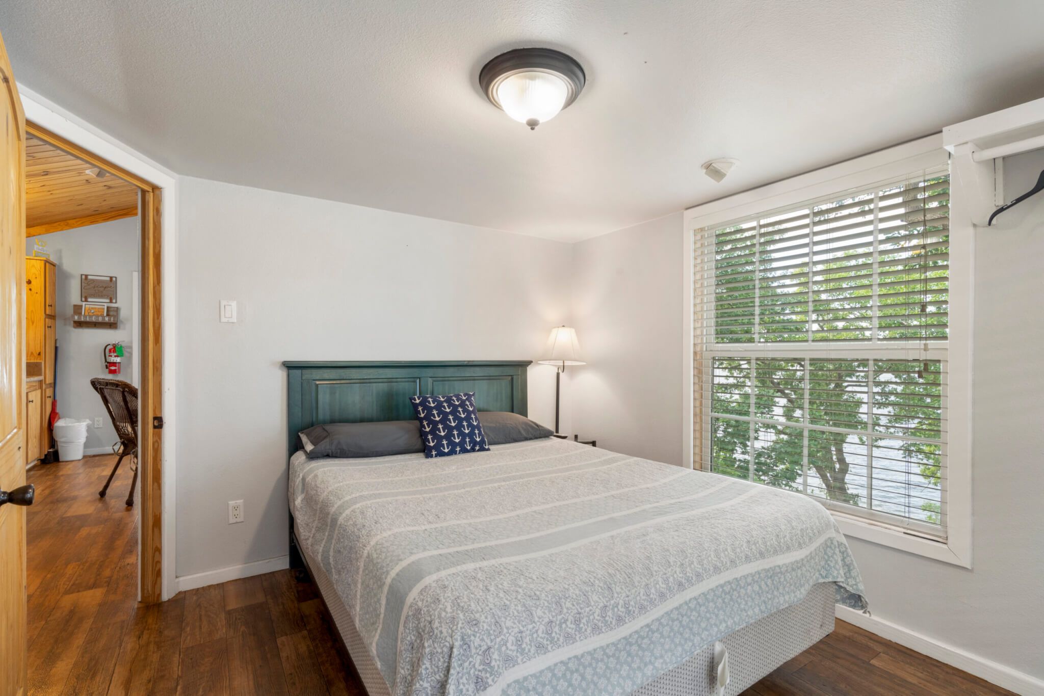 A bedroom with a bed, a nightstand, a lamp, a window with blinds, and a door leading to another room.