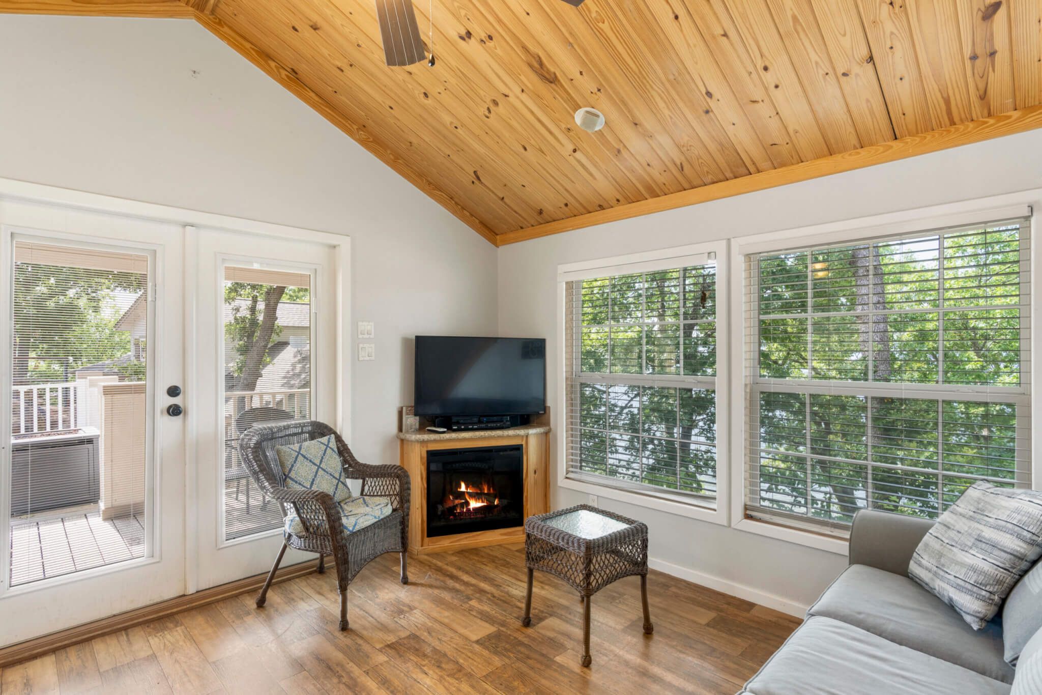 Una sala de estar acogedora con chimenea, televisor, mobiliario de mimbre y ventanas amplias que ofrecen vistas a árboles.