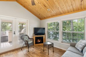 A cozy living room with a fireplace, a television, wicker furniture, and large windows offering a view of trees.