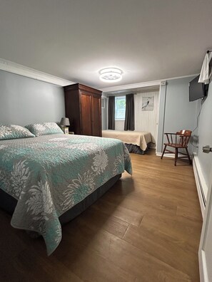 A bedroom with a large bed, a wooden wardrobe, a desk with a chair, and a television on the wall.