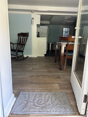An open doorway to a room with a wooden floor, a rocking chair, a dining table with chairs, a kitchen area with a refrigerator, and a window with blinds.