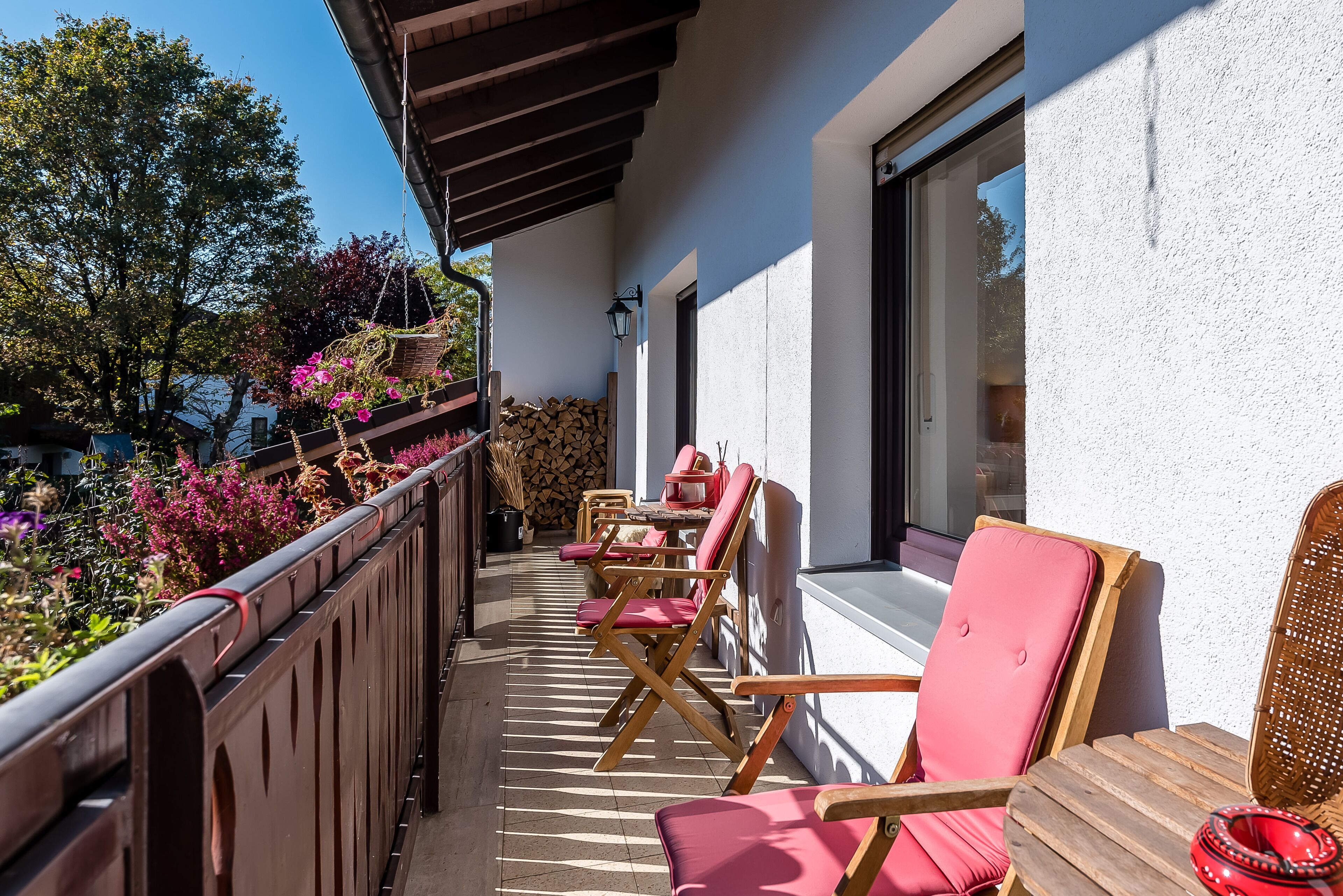 Ein Balkon mit Holzstühlen und einem Tisch, mit Blick auf einen Garten mit rosa Blumen und einem Holzstapel.