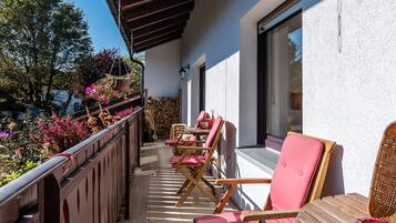 Ein Balkon mit Holzstühlen und einem Tisch, mit Blick auf einen Garten mit rosa Blumen und einem Holzstapel.