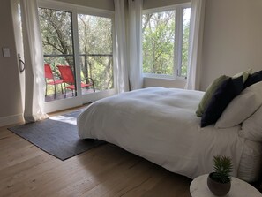 A bedroom with a large bed, a small round table with a plant, and a view of outdoor red chairs through a large window.