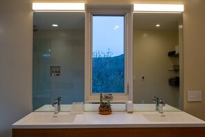 A bathroom with a large mirror, two sinks, a window with a view of trees, and a small plant on the counter.