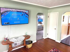 A living room with a flat-screen TV displaying a tennis match, a wooden side table, a doormat, and a hallway leading to a bedroom. - Nanny's Bungalow - Historic Downtown/Sanders Beach Location (Coeur d'Alene)