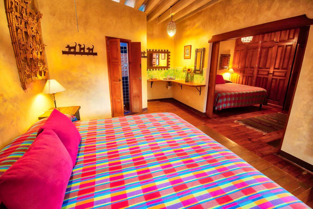 Une chambre à coucher avec une couvre-lit à carreaux colorés, une porte en bois, un miroir et une petite salle de bain avec un lavabo et un miroir.