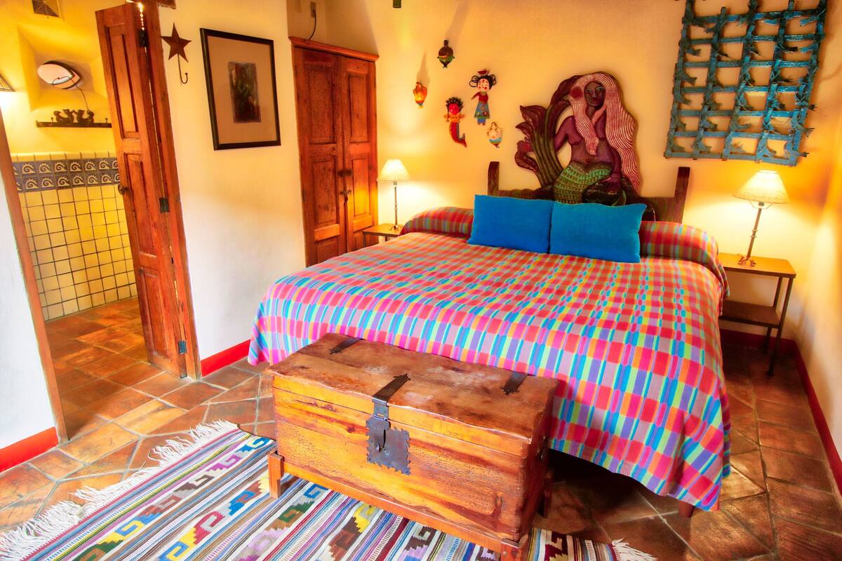 Une chambre avec une couvre-lit aux couleurs vives, une commode en bois et des œuvres d’art décoratives accrochées au mur.