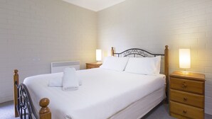 Un lit bien fait, avec du linge de lit blanc et une serviette pliée, une tête de lit en bois, deux tables de chevet avec des lampes et un radiateur au mur.