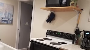 A kitchen with a white stove, a microwave on a wooden shelf, a coffee maker, and a painting of sailboats on the wall.